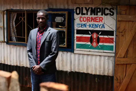 African man, a professional runner from Kenya, stands in front of his store selling athletic gear. A sign on the wall says "Olympics Corner Iten-Kenya", above a Kenyan flag.