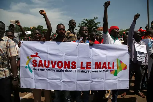 Several male Malian protestors, some with fists raised, hold a white banner with French writing translating as "Let's save Mali, All together hand in hand, let's save Mali."
