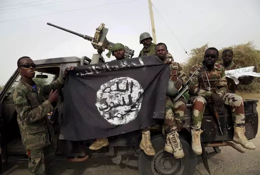 Six Nigerian soldiers, in an armored truck, hold up a flag seized from Boko Haram. The flag is black, with a white circle in the middle with black Arabic writing.