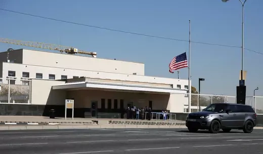 Gray car drives past pale concrete building flying U.S. flag at half mast. It is the U.S. Embassy in Johannesburg, South Africa.