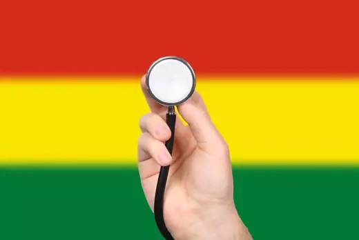A hand holds a stethoscope in front of a blurred Bolivian flag