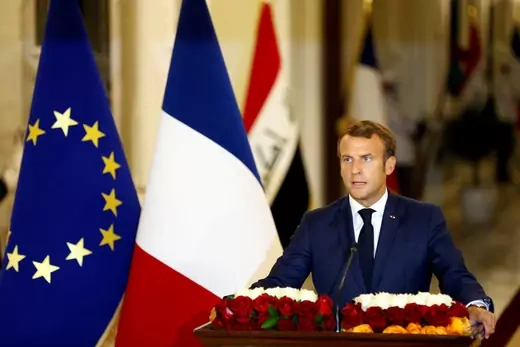 French President Emmanuel Macron delivers at a speech at a lectern covered with roses. He stands in front of a French flag, a European Union flag, and an Iraqi flag.