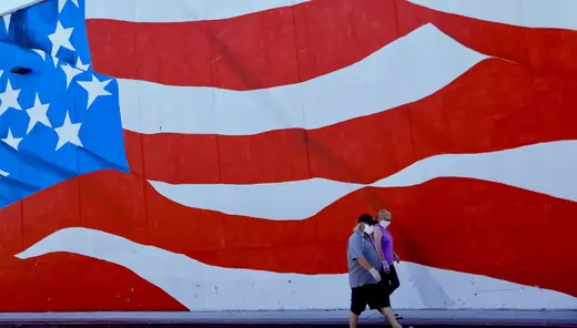 Two people in masks walk by a mural of the U.S. flag in Ocean Beach, California. 