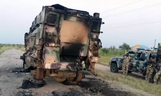 Burned out military vehicle, with tires removed, stands on a road in Nigeria, with soldiers looking on.