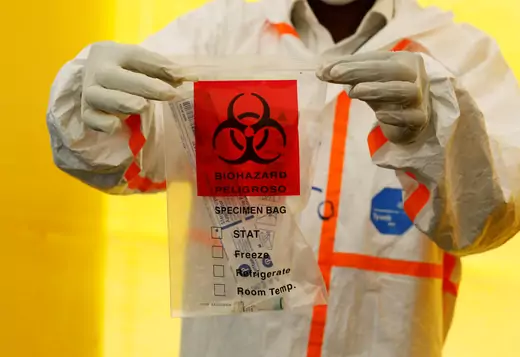 A health worker, dressed in white personal protective equipment (PPE), holds up a coronavirus testing kit. The front says "biohazard, peligroso."