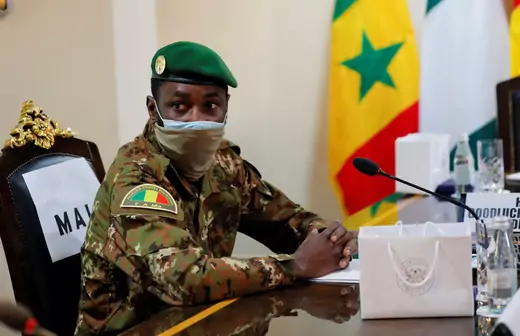 Malian coup leader Assimi Goita sits, in full military clothing, at a table during negotiations with ECOWAS. A red, yellow, and green flag is visible in the background.