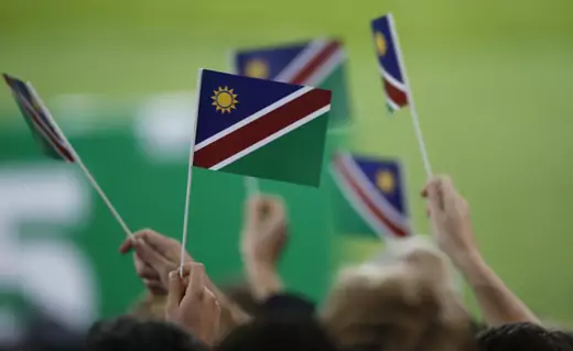 Small plastic flags of Namibia (a red stripe through blue and green corners, with a small yellow sun in the upper left blue corner) wave in a crowd.