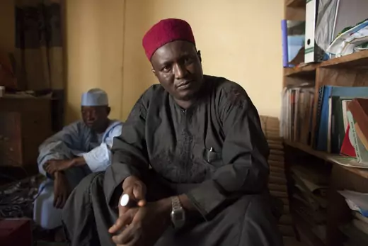 Nigerian man, wearing dark clothing and bright red hat, looks into the camera. A man sits behind, and a bookshelf lines the nearest wall.