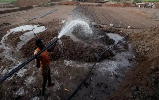 A man in a sleeveless shirt holds a pipe pouring water from the Nile river onto a patch of mud to make bricks on Khartoum, Sudan 