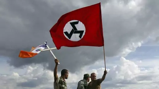 Three South African men hold flags of the Afrikaner Resistance Movement, including a red flag with three black sevens in the middle, and a blue, orange, and white flag pinned to a cross.