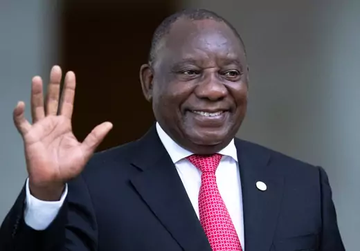 African man, South Africa's president Cyril Ramaphosa, waves and smiles. He is wearing a black suit, white shirt, and red tie.