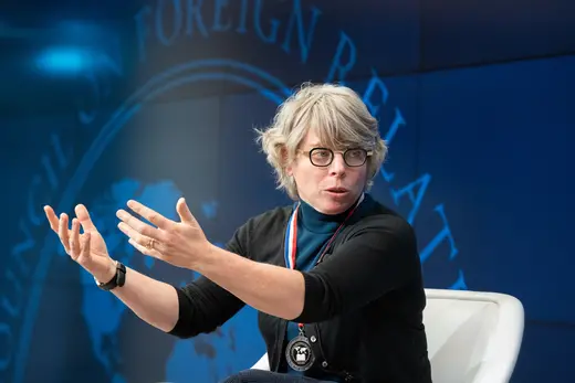 Jill Lepore, with short grey hair, wearing a blue turtleneck and black cardigan, black and gold glasses, and a medal around her neck, against a blue background