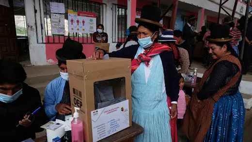 Women in skirts, aprons, and hats gather near a cardboard ballot box.  On of them, wearing a mask, is placing her ballot inside.