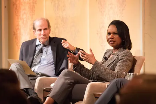 Michael Krasny and Condoleezza Rice sit in chairs in front of a gold background.