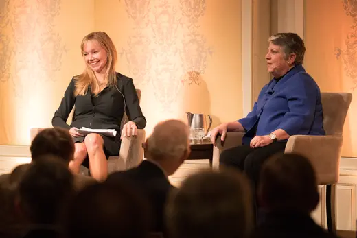 Two panelists sit in front of a gold patterned wall