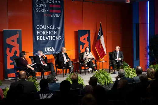 Five panelists sit on stage in front of an orange wall with blue banners