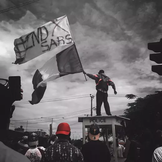 A man standing on top of a wooden structure with the word "police" painted on. He is waving two flags--a Nigerian national flag and one with #EndSARS spray-painted on it. The picture is mostly black and white.