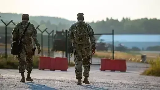 An image from behind of two men dressed in camouflage fatigues. They are walking across gravel near a fence and red road barriers. Trees appear in the distance.
