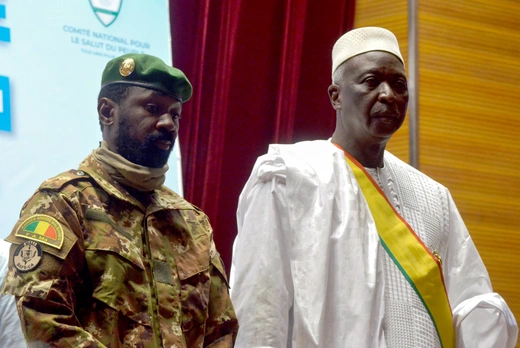 Two African men, one in military clothing, one in traditional white clothing, attend a presidential inauguration. The flag of Mali can be seen.