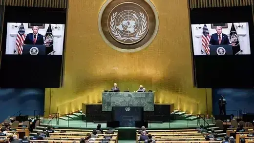 People sitting spaced apart at long tables watching a speech by Trump, which is shown on two screens that flank a podium at the UN headquarters.
