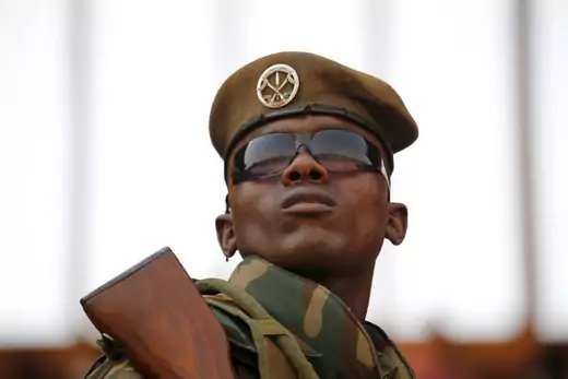 Nigerien soldier, wearing fatigues, sunglasses, a brown hat, and with a gun resting on his shoulder, stands guard.
