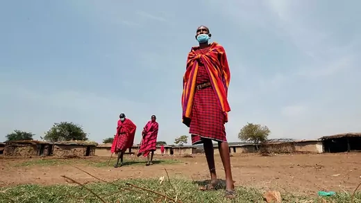 Three Maasai elders, wearing traditional costumes in red and gold, with face masks.