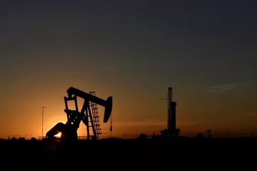 A pump jack operates in front of a drilling rig at sunset in an oil field in Midland, Texas U.S.