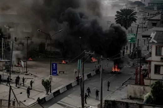 A picture of fires burning on the streets of Lagos, Nigeria. Thick black smoke is billowing into the sky. Several armed men can be seen patrolling the road.