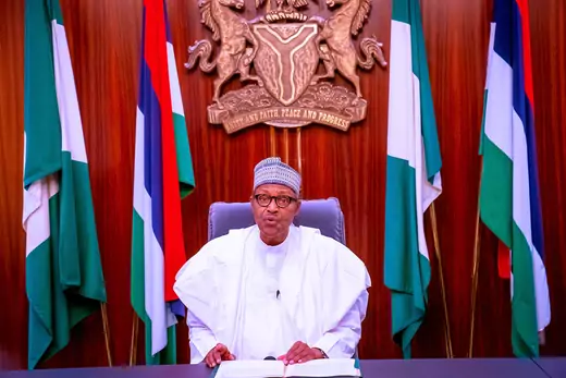 Nigerian President Muhammadu Buhari can be seen wearing traditional clothing before giving a televised address. Nigerian flags and another flag can be seen behind the president, along with the Nigerian coat of arms.