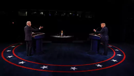 Joe Biden and Donald Trump gesture at each other from behind their podiums while Kristen Welker is seated between them on the debate stage. 
