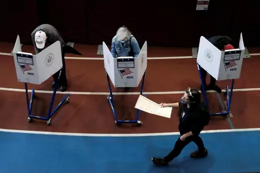 Three people stand behind privacy screens to cast their vote while another walks in front of them carrying a ballot to a voting booth. 