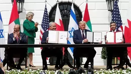 Four men in suits sitting at a long table, with three of them holding up signed agreements.  The other man and a woman behind them are clapping.  Flags of the United States, Bahrain, Israel, and the United Arab Emirates are in the background.