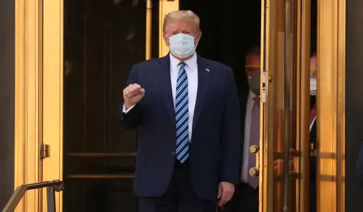 President Donald Trump, wearing a dark blue suit and blue tie, makes a fist as he leaves Walter Reed hospital. He is wearing a blue mask.
