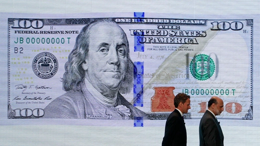 Two men in suits walk by a large US 100 bill on the wall at the Department of the Treasury