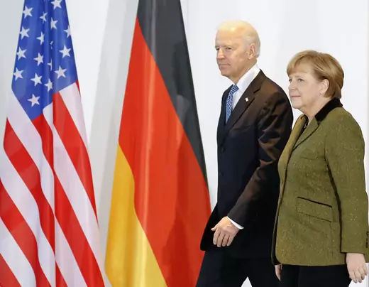 Joe Biden walks beside Angela Merkel in front of U.S. and German flags.