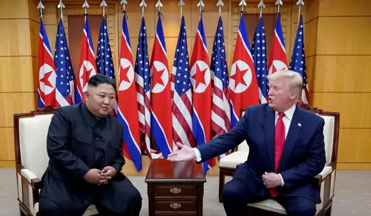 U.S. President Donald J. Trump reaches his hand out to North Korean leader Kim Jong Un while sitting down in front of several United States and North Korean flags at the demilitarized zone separating the two Koreas, 