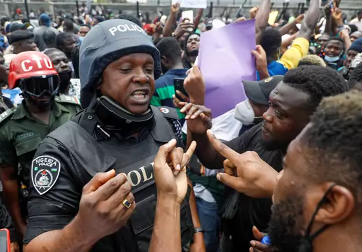 A Nigerian police officer in full uniform speaks with two protesters during protests against the Special Anti-Robbery Squad (SARS). There is a large crown in the background.