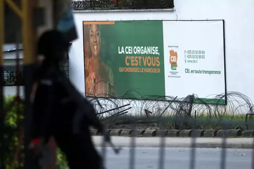 A soldier can be seen leaning against a post with an assault rifle. A billboard for Ivory Coast's Independent Electoral Commission is seen in the background.