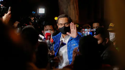 Reporters crowd around former Peruvian President Martin Vizcarra, who is wearing a blue jacket and mask and holding up his hand.  It is dark outside.