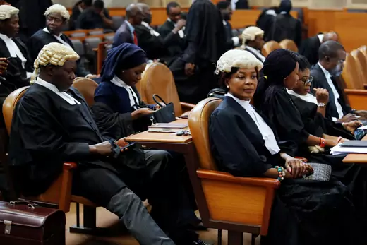 A group of Nigerian lawyers are seen sitting in chairs awaiting a trial. Several are wearing wigs that resemble those used during the British colonial period.