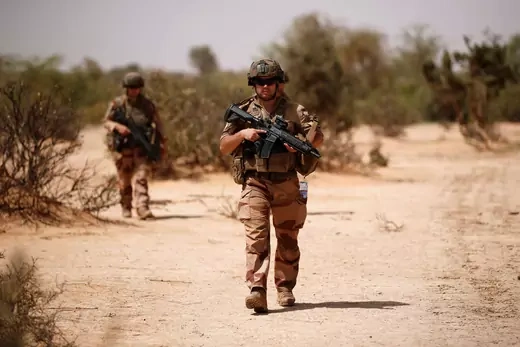 Two French soldiers are seen walking through an arid area in Mali. Both are wearing desert camouflage, military equipment, and assault rifles.
