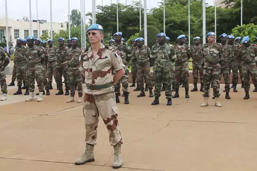 A group of UN peacekeepers standing at attention as they prepare to begin a mission in Mali in 2013. All are wearing blue UN berets. One man wearing desert fatigues stands in the front, with a group of soldiers wearing darker camouflage standing behind him.