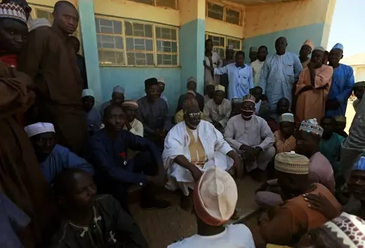 A group of parents are seen gathered outside of a school in northern Nigeria. Many are wearing traditional Islamic dress. Some parents are sitting, others standing.