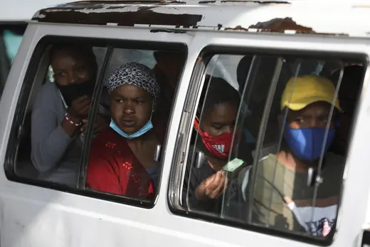 A group of South African commuters can be seen sitting inside of a transport van. Some are wearing masks, while others are not.