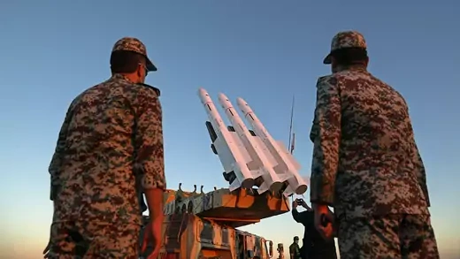 Two Iranian soldiers with their backs to the camera frame four missiles poised to launch in the background