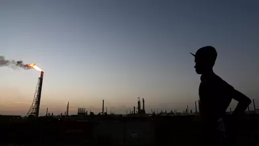 A silhouetted person stands in the foreground with industrial structures, including a tower with smoke and fire coming out of it, in the background. The photo appears to have been taken at either dusk or dawn. 