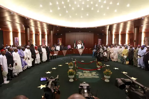 Nigeria's President Muhammadu Buhari swears in ministers into his cabinet in Abuja, Nigeria on November 11, 2015. The room is well-lit with a green carpeted floor, with the Nigerian coat of arms surrounded by four pots of flowers. The ministers are standing around the room, while Buhari is seated at the head.