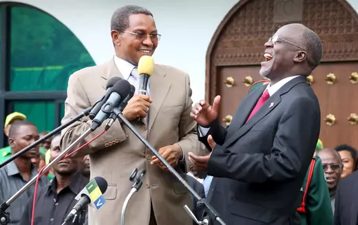 Tanzanian President John Magufuli and his predecessor, Jakaya Kikwete, can be seen speaking to one another ahead of a speech for the ruling party, the Chama cha Mapinduzi. Kikwete is holding a microphone while Magufuli leans back laughing.