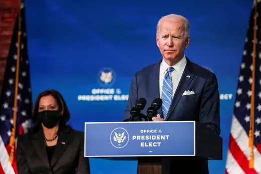 Vice President-Elect Kamala Harris sits behind President-Elect Joe Biden as he speaks at a podium labelled "Office of the President Elect." 