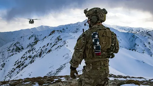 A U.S. Air Force member stands atop a snowy mountain in Afghanistan. A helicopter is seen in the distance.